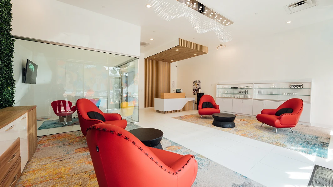 A modern, bright waiting area with white tile floors, featuring four curved red chairs with black pillows arranged around two round black coffee tables. One wall is covered in green artificial foliage with a white neon sign that says "Vibrant Vibes." There is a glass-walled conference room in the center and a reception desk with a vertical wood slat background on the right.