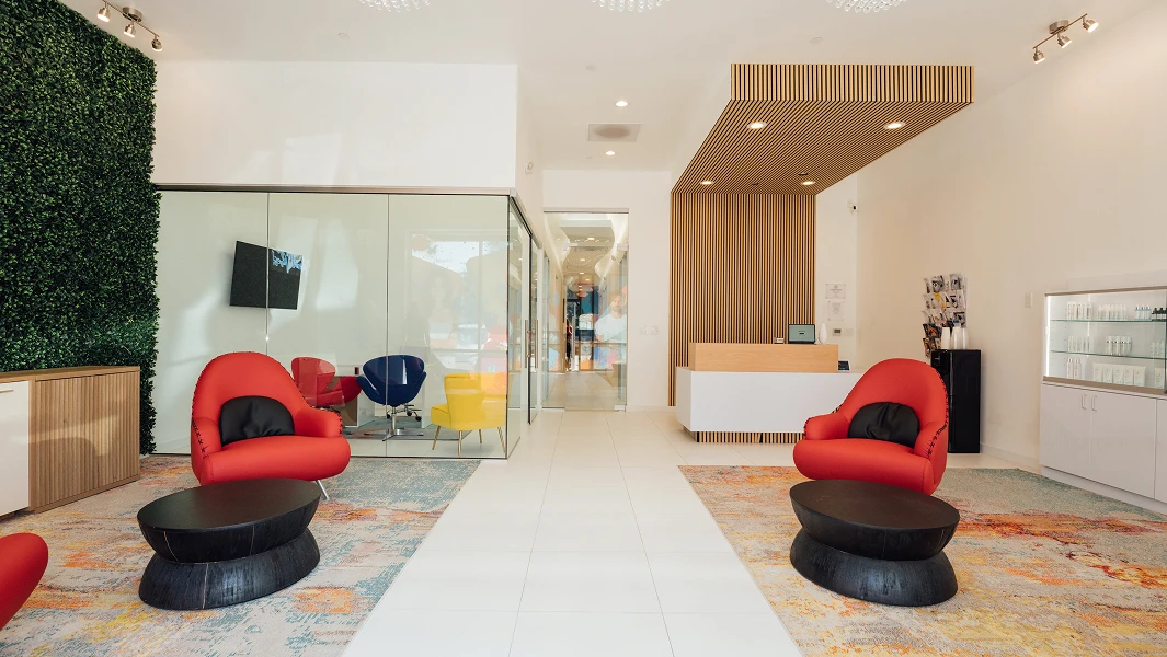 A modern, bright waiting area with white tile floors, featuring four curved red chairs with black pillows arranged around two round black coffee tables. One wall is covered in green artificial foliage with a white neon sign that says "Vibrant Vibes." There is a glass-walled conference room in the center and a reception desk with a vertical wood slat background on the right.