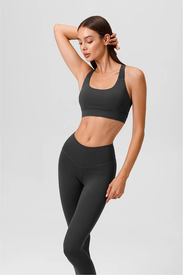 A woman with long dark hair, wearing a black sports bra and matching high-waisted leggings, strikes a confident pose with one hand touching her head and her body angled against a plain light gray background. - Procedures in The Colony, TX