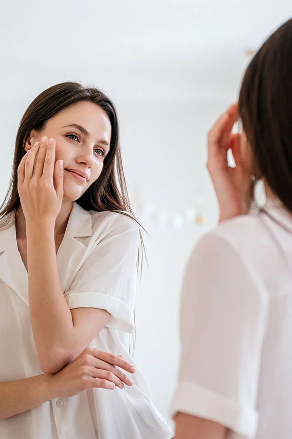 A young woman with long dark hair, wearing a white shirt, is looking into a mirror and gently touching her cheek with one hand, smiling faintly at her reflection. - BBL Forever Young Laser in The Colony, TX