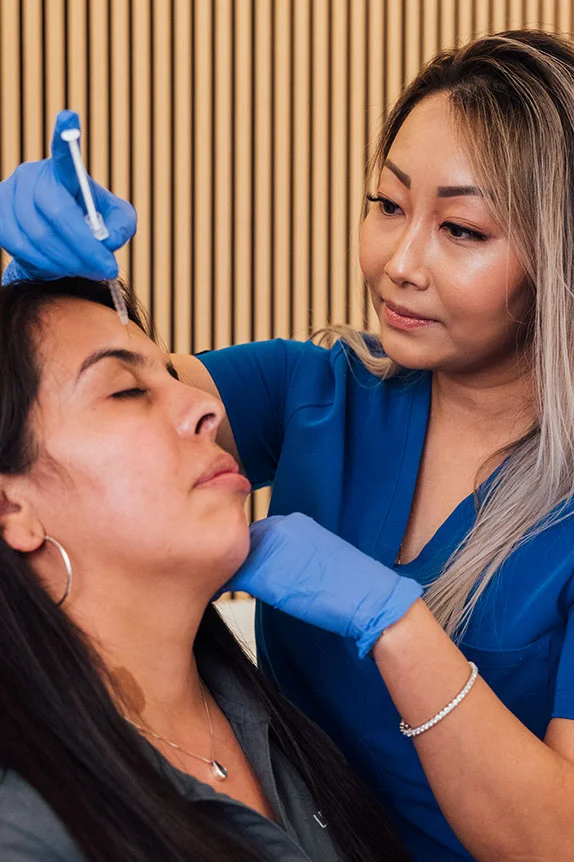 A female practitioner in blue scrubs and blue gloves is administering an injection, likely Botox or a filler, to a patient's forehead. The patient is lying back with her eyes closed. The background has a textured wall of vertical wooden slats. - Botox and Wrinkle Relaxers in The Colony, TX