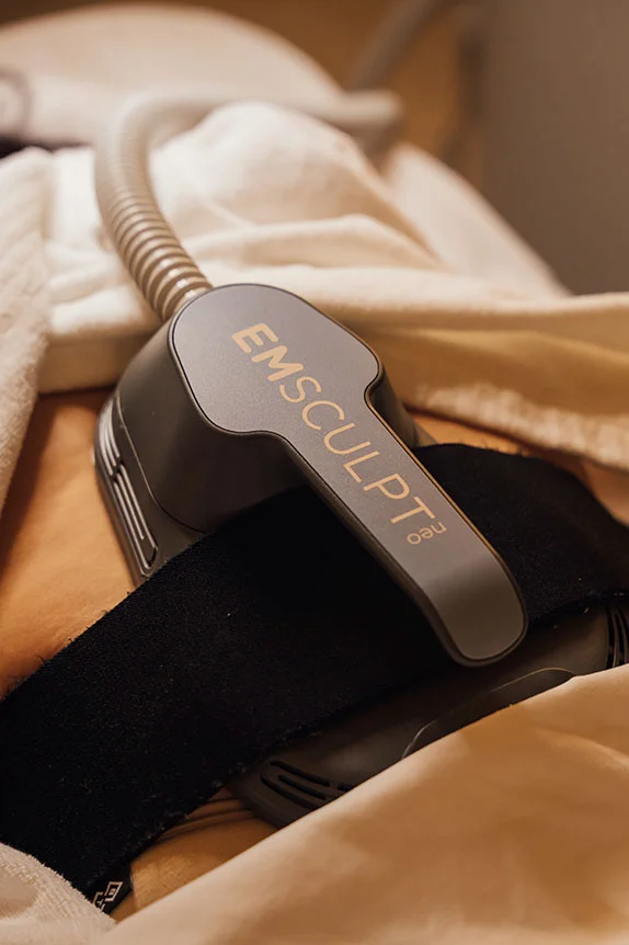 A close-up view of the EM Sculpt Neo applicator device placed on a patient's abdomen, secured with a black strap, with a white towel visible around the area. - Emsculpt Neo in The Colony, TX