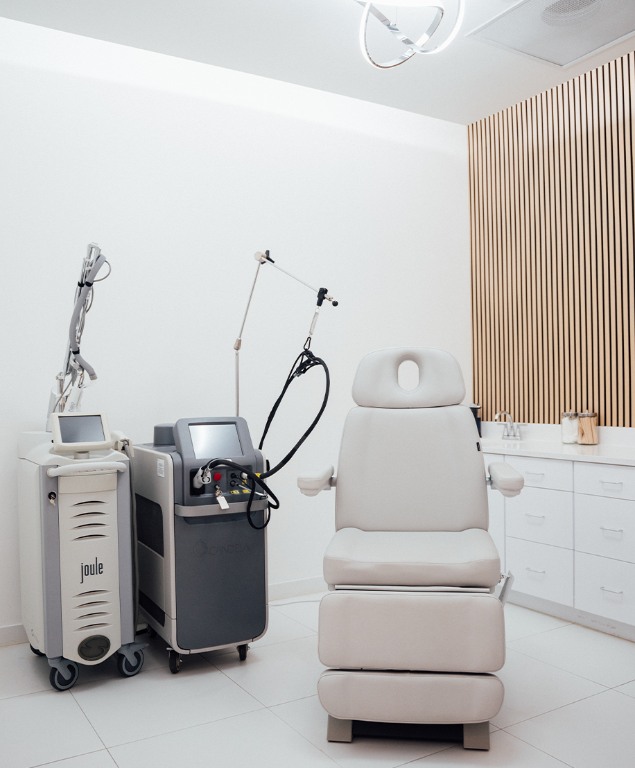 A clean, modern treatment room with white walls and tile floor. The room contains a light gray adjustable treatment chair, two medical laser machines, white cabinets, and a wall accent panel made of vertical wooden slats.