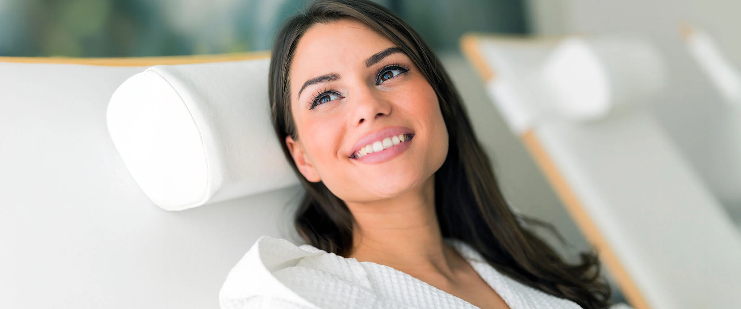 A smiling woman with long dark hair is comfortably seated in a reclining chair, wearing a white robe and looking up and to the side. The chair has a white headrest. - NAD Therapy in The Colony, TX