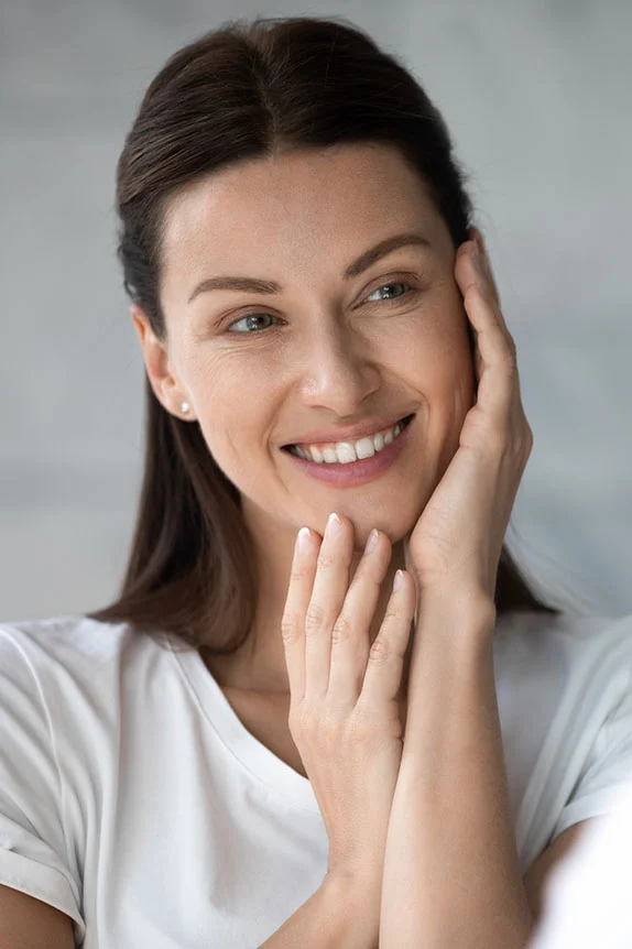 A young woman with long, dark hair looks directly at the camera, gently touching her cheeks with both hands and smiling. She is wearing a white t-shirt, and the background is blurred, showing a light-colored, possibly tiled, surface. - ProCell Microchanneling in The Colony, TX