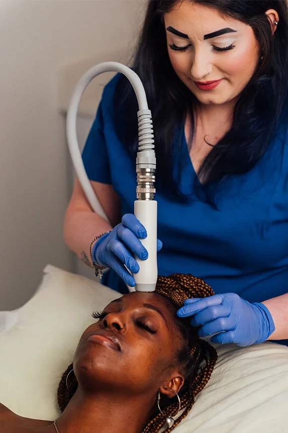 A female technician in blue scrubs and blue gloves performs an RF microneedling treatment on a patient lying on a treatment bed. The technician is holding a handheld device near the patient's forehead. - RF Microneedling Vivace in The Colony, TX